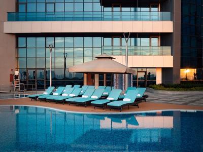 Piscina a sfioro con sdraio e ombrellone davanti a un edificio moderno con facciata in vetro.