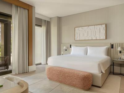 Modernes Schlafzimmer mit großem weißem Bett, beigem Hocker, Nachttischen und bodentiefen Fenstern.
