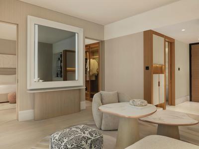 Moderner Hotelraum mit Sitzbereich, Tisch, Spiegel und Blick ins Schlafzimmer, helle, warme Farbtöne.