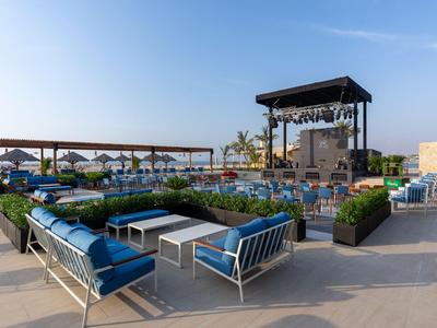 Nowoczesny lounge na zewnątrz z niebieskimi siedzeniami i sceną w słonecznym otoczeniu hotelu.