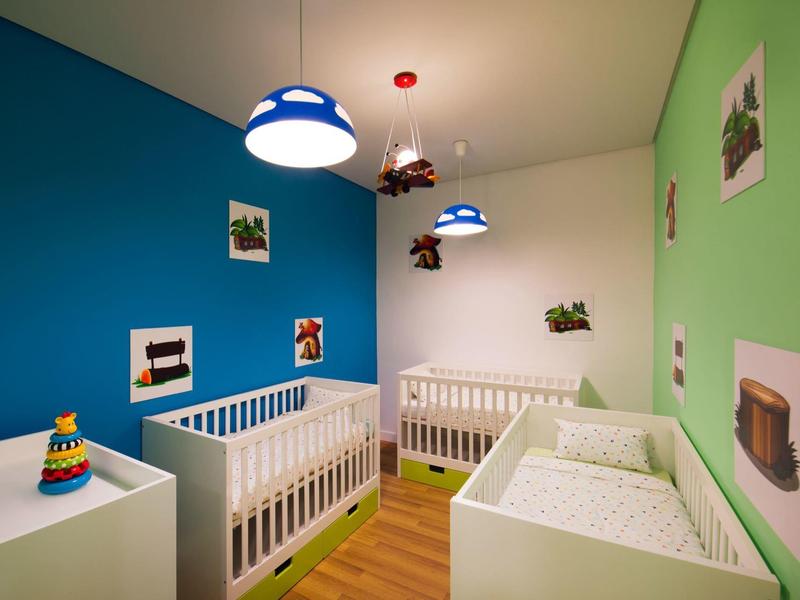 Hell eingerichtetes Babyzimmer mit blauen und grünen Wänden, zwei Kinderbetten und bunten Lampen.