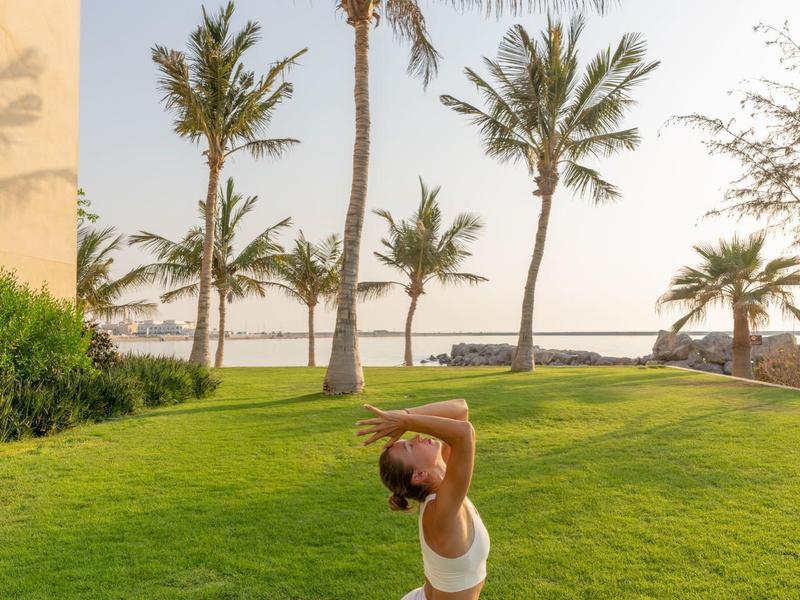 Frau macht Yoga im Spagat auf einer Matte mit Palmen und Meer im Hintergrund.