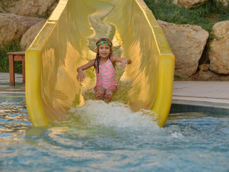 Ein Kind rutscht auf einer gelben Wasserrutsche ins blaue Schwimmbecken, umgeben von Felsen.