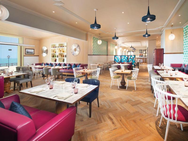 Helles Restaurant mit bunten Sesseln, Holzboden und großen Fenstern für viel Tageslicht.