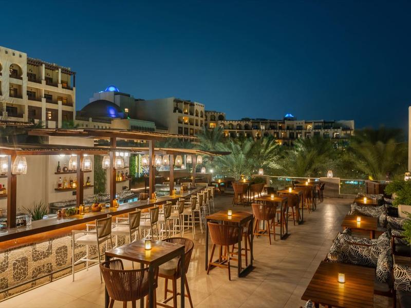 Freiluftrestaurant mit Holztischen, Stühlen und warmen Lichtern in einer nächtlichen Stadtlandschaft.