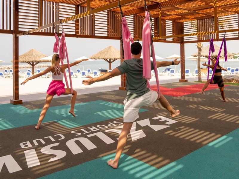 Mehrere Personen üben im Fitnessclub mit pinken Lufttüchern unter einem Holzdach in Strandnähe.