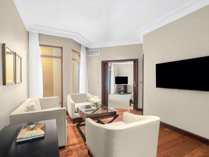 Helles Wohnzimmer mit weißen Sesseln, Holzboden, Wand-TV und Fenster mit Blick ins Nachbarzimmer.