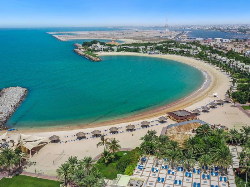Breiter Sandstrand mit klarem blauen Wasser, grün bewachsene Uferzone und Stadt im Hintergrund.