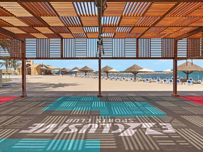 Blick durch Holzpergola auf Sandstrand mit Sonnenschirmen und ruhigem blauem Meer.