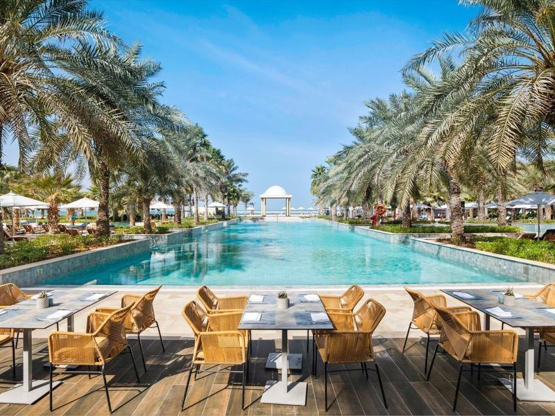 Ein großer Pool mit Palmen und Holzstühlen, blauer Himmel und weißen Sonnenschirmen.
