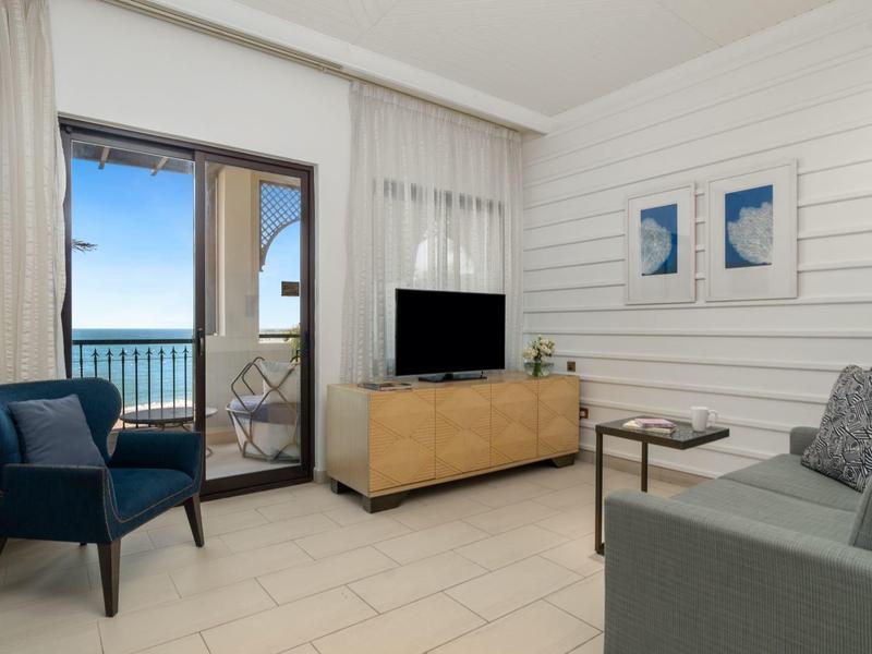 Helles Wohnzimmer mit grauem Sofa, blauem Sessel, TV, Holzmöbel und Blick auf das Meer.