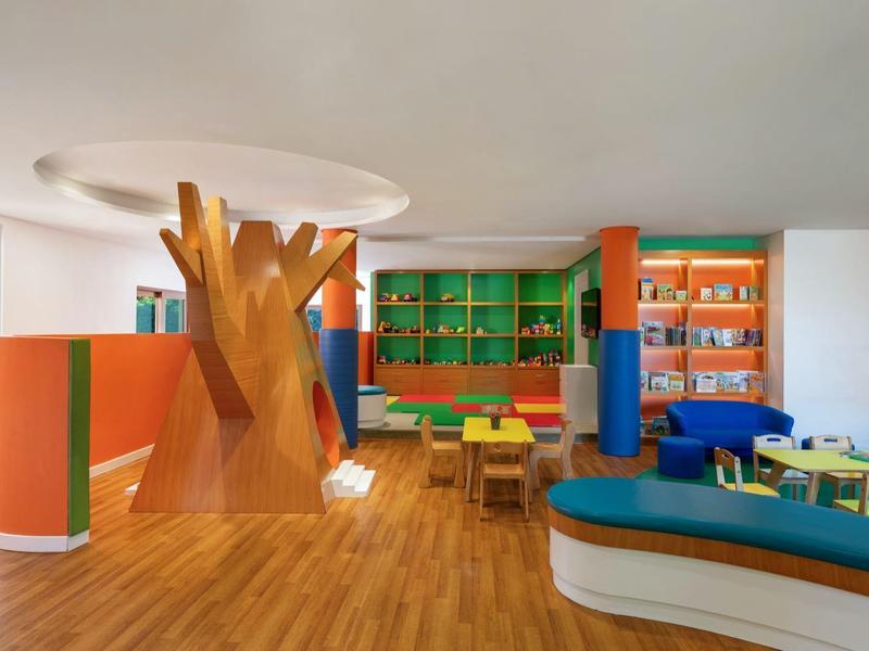 Bunter Raum mit Baum-Skulptur, Holzboden, Tischen und Regalen voller Bücher in Kinderbibliothek.