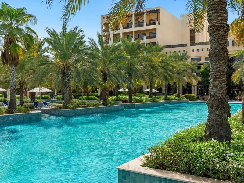 Ein Hotel mit großem Pool umgeben von Palmen und Sonnenschirmen in tropischer Umgebung.