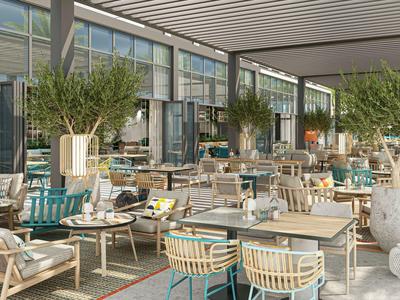 Světla restaurace se dřevěnými židlemi, stoly a velkými rostlinami pod moderní skleněnou střechou.