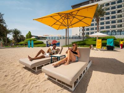 Dva lidé odpočívají na lehátkách pod velkým žlutým slunečníkem na pláži před hotelem.