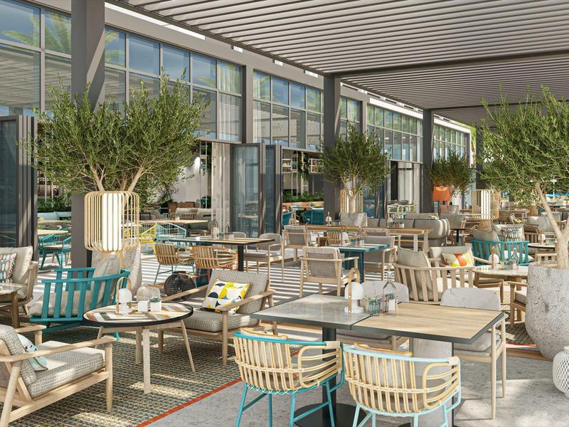 Světla restaurace se dřevěnými židlemi, stoly a velkými rostlinami pod moderní skleněnou střechou.