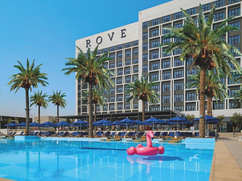 Moderní hotel s velkým bazénem, palmami a nafukovacím růžovým plameňákem ve vodě.