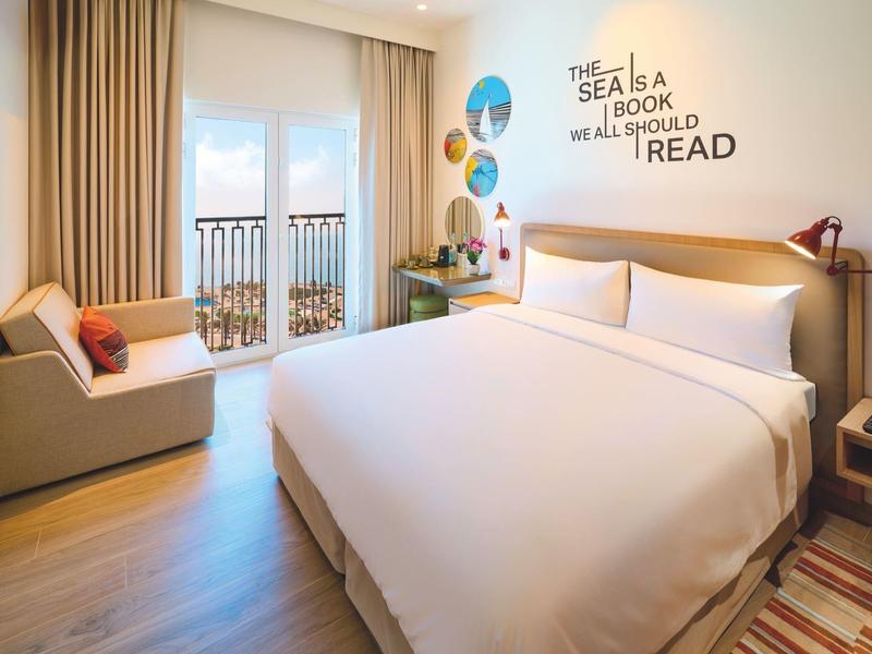 Světle zařízený hotelový pokoj s manželským lůžkem, pohovkou a balkónem s výhledem ven.