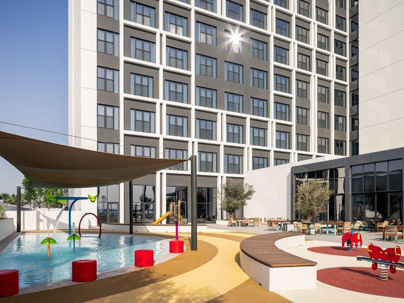 Moderní hotelová venkovní plocha s dětským hřištěm, bazénem a sluneční clonou za jasné oblohy.