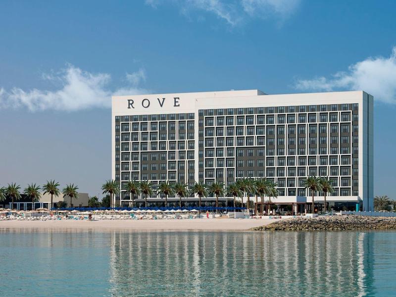 Moderní hotelová budova u vody s jasnou oblohou a palmami v popředí.