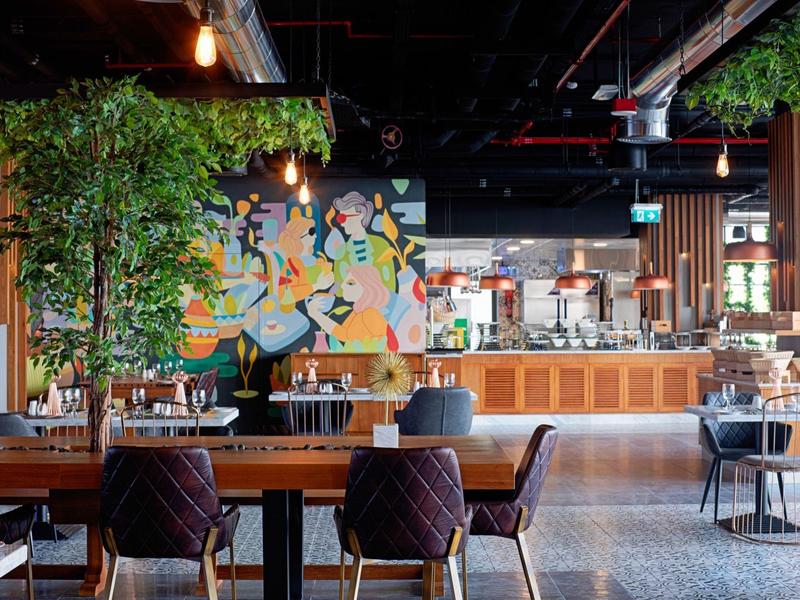 Moderner Gastraum mit Holztischen, Pflanzen, buntem Wandbild und offener Küchentheke.