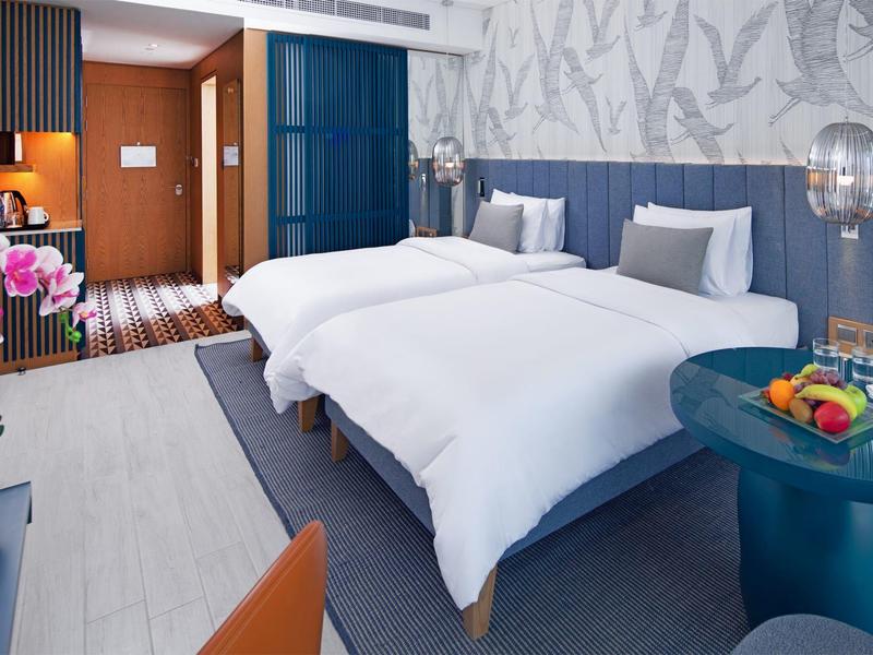 Modernes Hotelzimmer mit zwei Betten, blau-grauen Wänden und einem runden Tisch mit Obst.