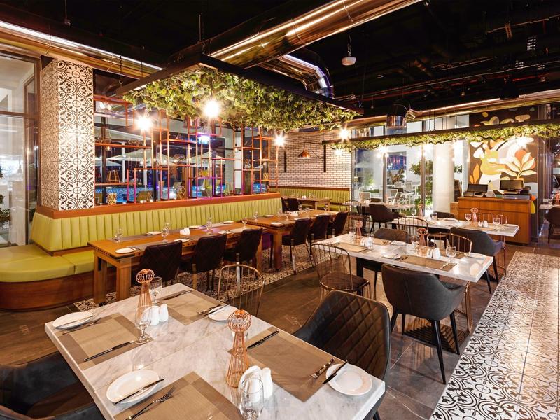 Modern gestaltetes Restaurant mit Holz- und Metallmöbeln, gelben Sitzbänken und grünen Pflanzen.