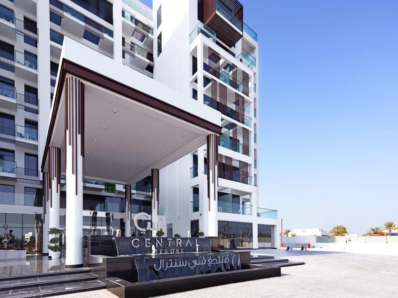 Moderner Gebäudeeingang mit Glasfassade, Säulen und lichtdurchflutetem blauen Himmel im Hintergrund.