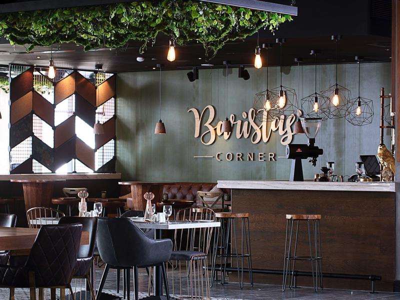 Moderner Café-Innenraum mit Bar, Metallstühlen, Holz- und grünen Designelementen, warmem Licht.