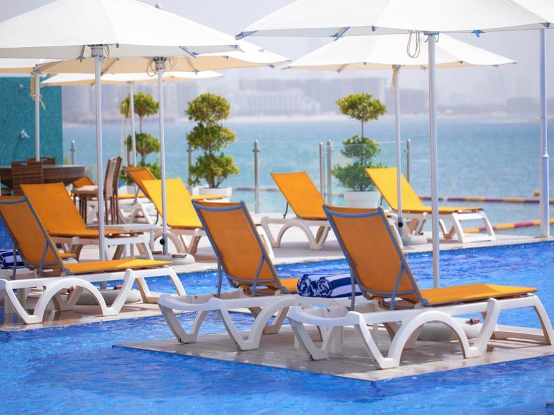 Leeres Schwimmbad mit orangenen Liegestühlen und weißen Sonnenschirmen am blauen Wasser.