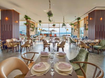 Hell eingerichtetes Restaurant mit Holz- und Pflanzenakzenten, rundem Mobiliar und großem Fensterblick.