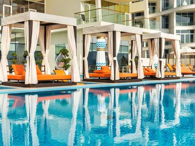 Pool mit blauer Wasserfläche, weißen Sonnendächern und orangefarbenen Liegen vor Hotelgebäude.