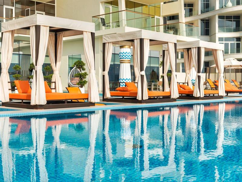 Pool mit blauer Wasserfläche, weißen Sonnendächern und orangefarbenen Liegen vor Hotelgebäude.