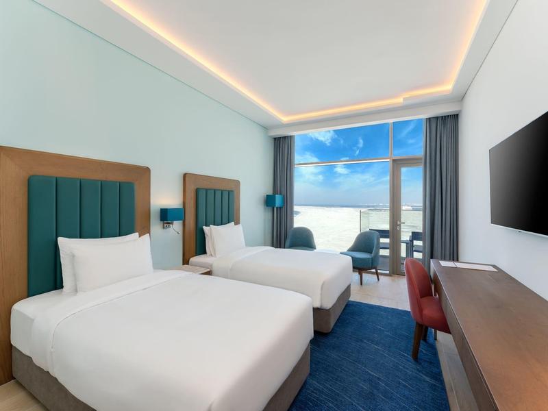 Habitación de hotel moderna con dos camas, alfombra azul, escritorio y vista al mar a través del ventanal.