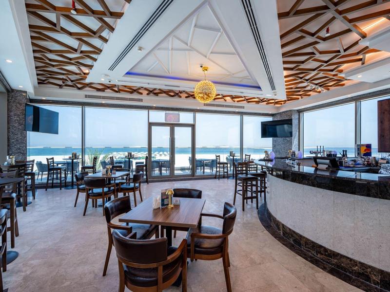 Restaurante moderno de hotel con mesas y sillas de madera, barra y grandes ventanas con vista al mar.