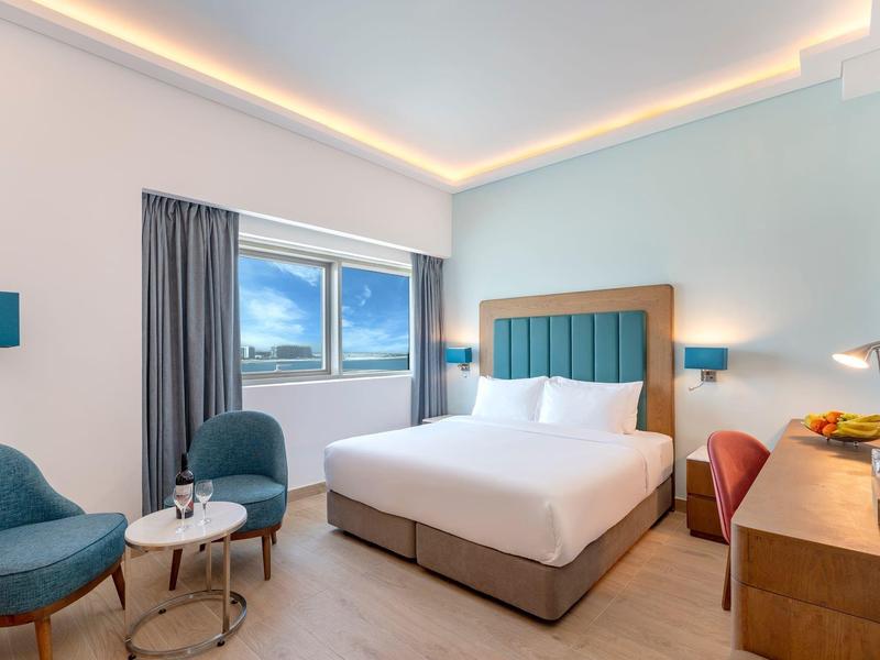 Habitación de hotel moderna con cama doble, dos sillones, ventana grande y escritorio con televisor.