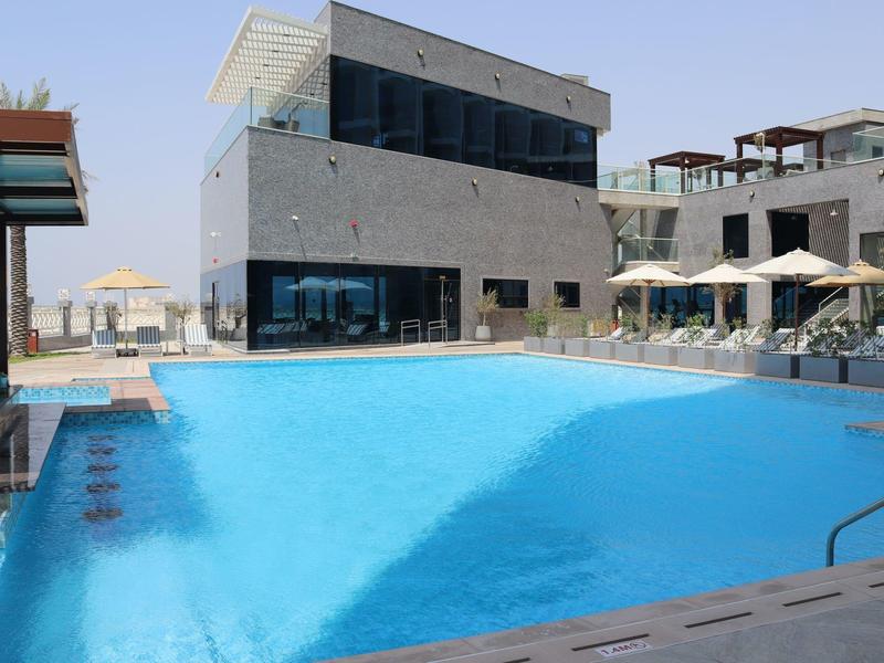 Piscina moderna de hotel con agua azul clara, sombrillas y edificio de varios pisos al fondo.
