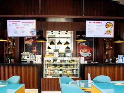 Nowoczesna kawiarnia z niebieskimi krzesłami, drewnianymi ścianami i cyfrowym menu nad ladą.