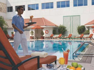 Pracownik hotelu niosący tacę obok basenu z leżakami i napojami.