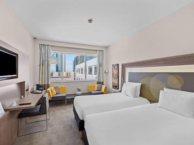 Modern hotel room with two single beds, desk, TV, and city view.