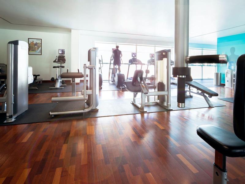 Gym with equipment and wooden floor, illuminated by large windows.