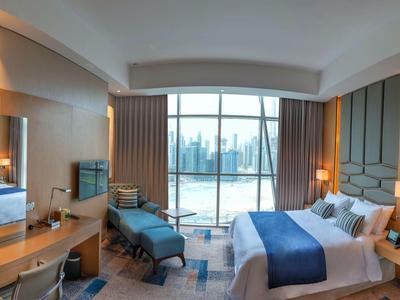 Modern hotel room with large bed, desk, and balcony overlooking a cityscape.