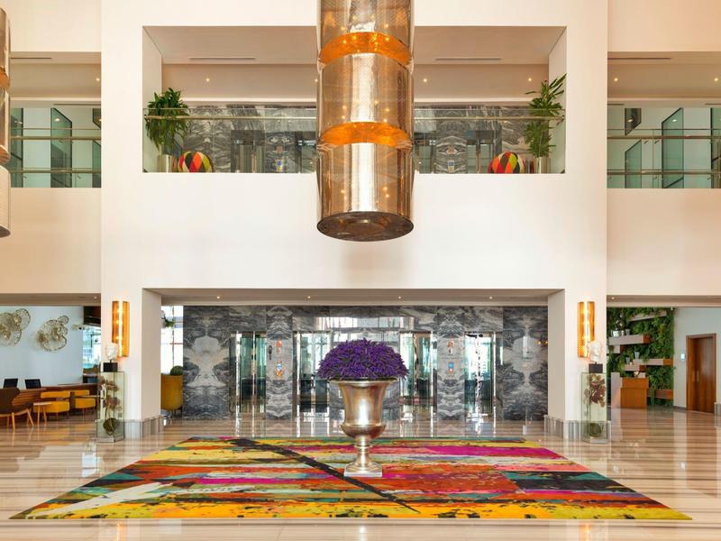 Bright hotel lobby with large colorful rug and central flower arrangement.