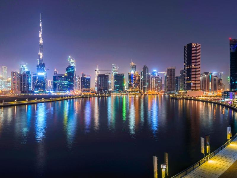 Nighttime skyline of a modern city with illuminated skyscrapers and water reflections.
