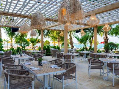 Überdachter Außenbereich eines Restaurants mit Tischen, Stühlen und Blick auf tropische Pflanzen und Wasser.