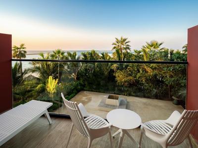 Balkon mit weißen Stühlen und Tisch, Blick auf Palmen und Meer bei Sonnenuntergang.