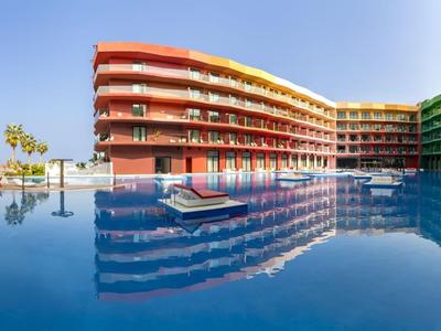 Moderner Hotelpool mit lichtdurchflutetem Gebäude und klar blauem Wasser unter klarem Himmel.