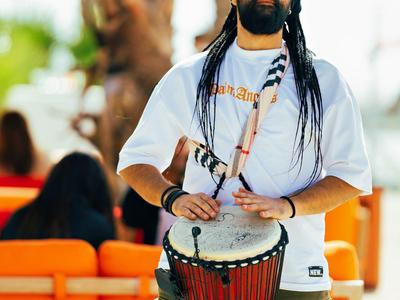Mann mit Dreadlocks und Sonnenbrille spielt rote Trommel draußen vor orangefarbener Couch.