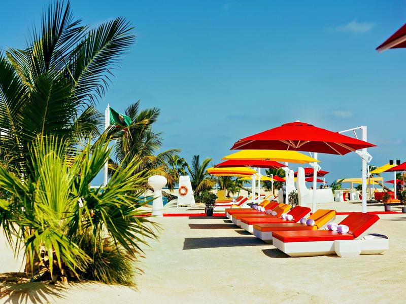 Beach with sunbeds and red umbrellas under blue sky and palm trees.