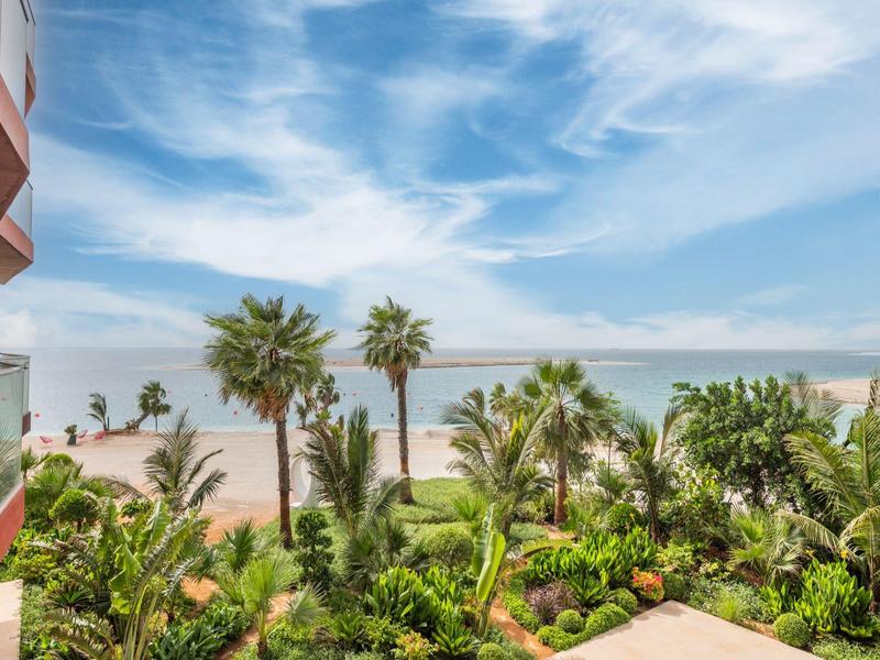 View from a balcony of palm trees and sandy beach under a blue sky.